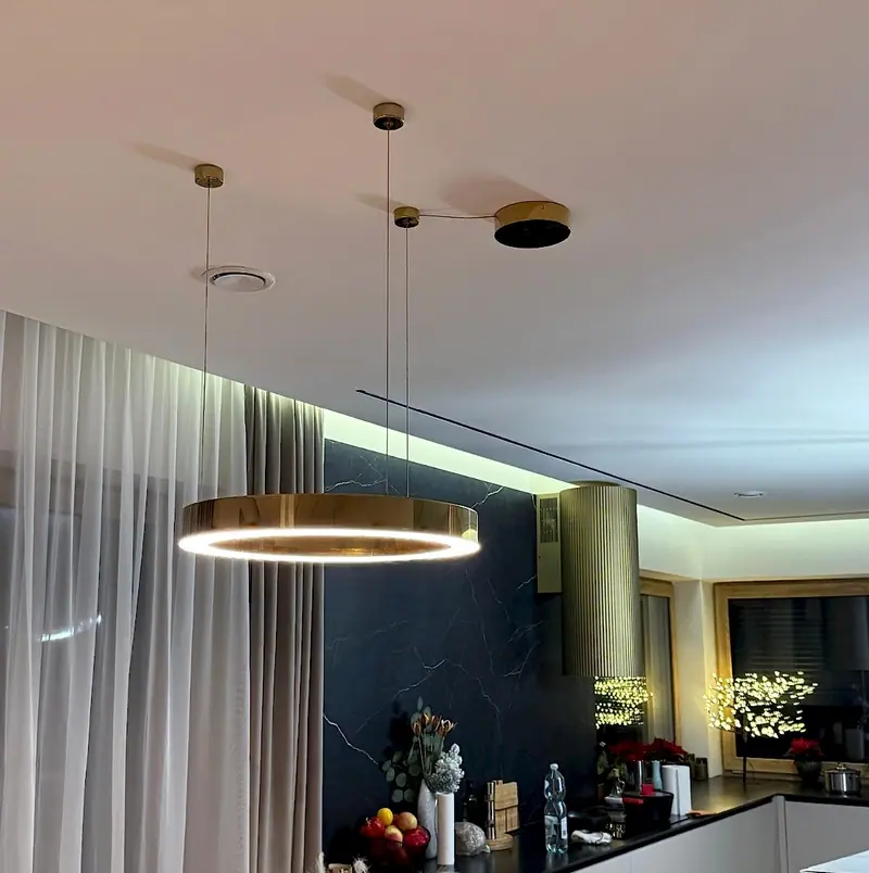Złota lampa wisząca w kształcie pierścienia LED nad stołem, w tle kuchnia z czarnym blatem i ukrytą taśmą LED pod sufitem
